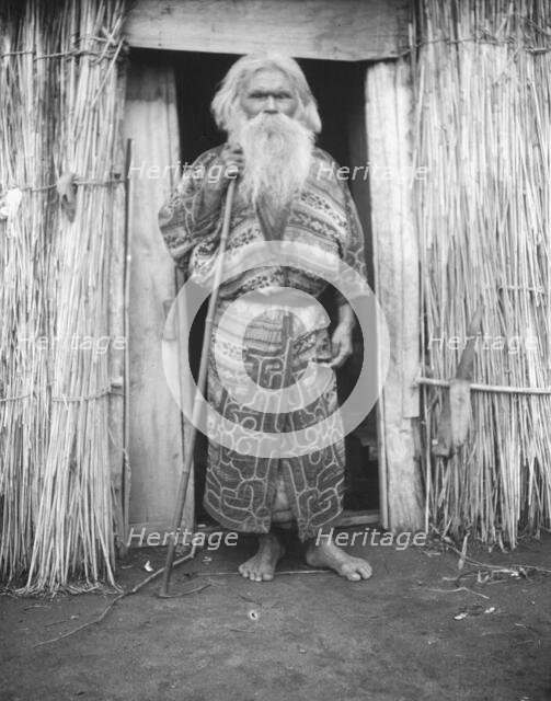 Ainu man holding a staff standing at the doorway of a hut, 1908. Creator: Arnold Genthe.