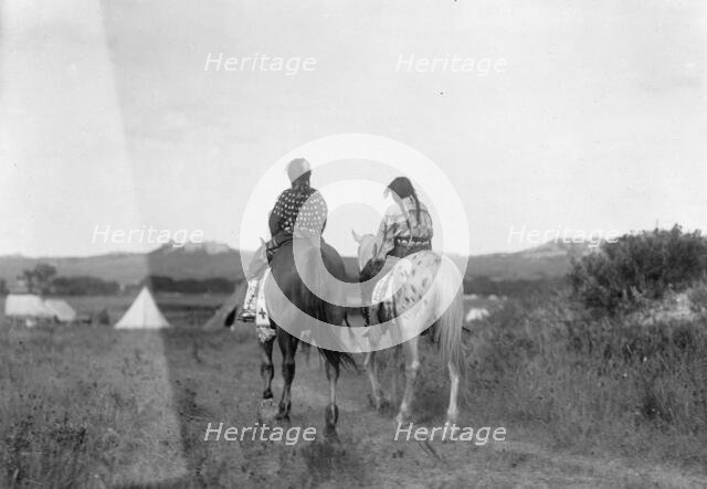 Two daughters of a chief on horseback, riding away from camera toward tents in background, c1907. Creator: Edward Sheriff Curtis.