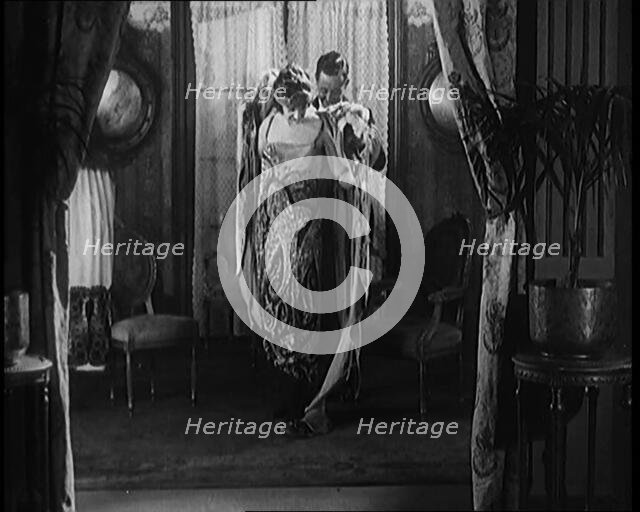 A Male Civilian Helping a Female Civilian Remove Her Fur Coat Revealing an Ornated Gown, 1920. Creator: British Pathe Ltd.