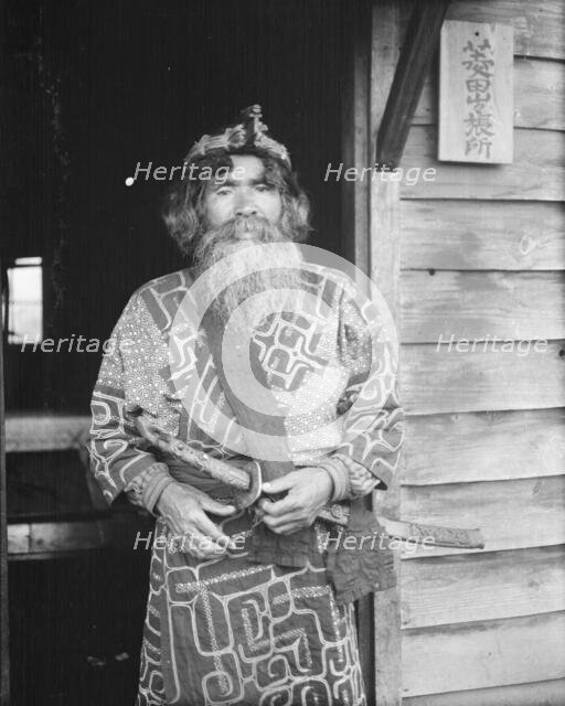 Ainu chief wearing a headdress and holding a sword standing in a doorway, 1908. Creator: Arnold Genthe.