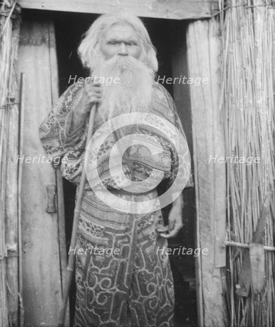 Ainu man holding a staff standing at the doorway of a hut, 1908. Creator: Arnold Genthe.
