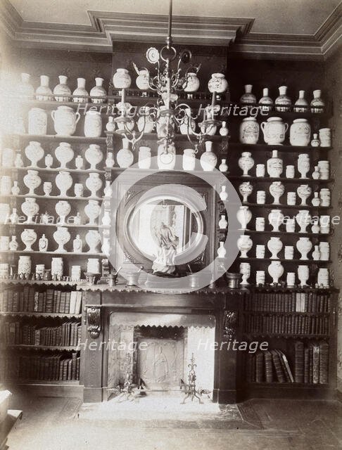 Drug jars and books on shelves surrounding a mantlepiece with a statue and a mirror, c1900.. Creator: Unknown.