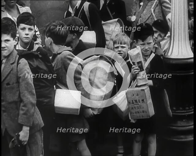 British Child Evacuees Standing on a Pavement With Bags and Boxes as Adults are Standing..., 1939. Creator: British Pathe Ltd.