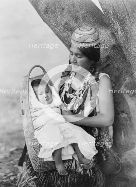 Hupa mother, three-quarter length portrait, seated, facing left, holding baby, c1923. Creator: Edward Sheriff Curtis.