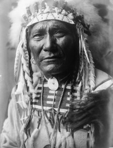Ghost Bear, Crow Indian, Montana, head-and-shoulders portrait, facing front, feather..., c1908. Creator: Edward Sheriff Curtis.