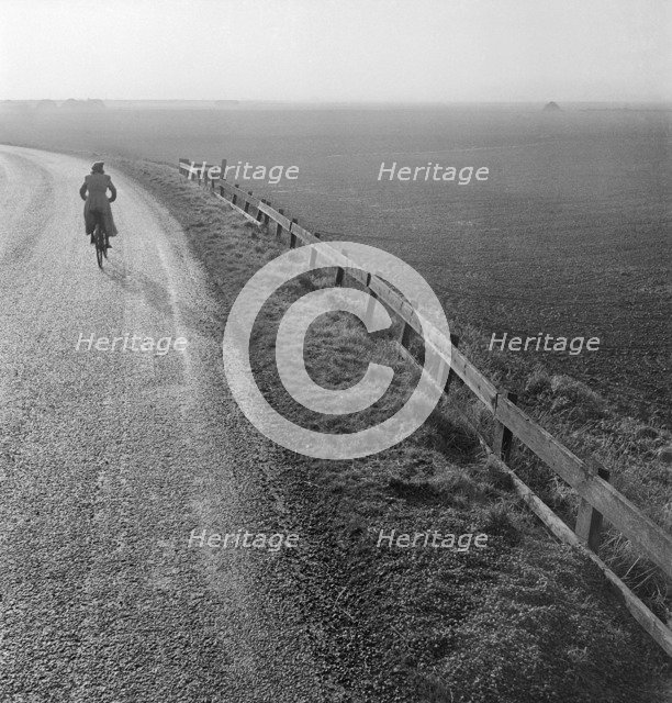 Woman cycling along a road raised up above the surrounding fenland, Cambridgeshire, early 1950s. Artist: John Gay.