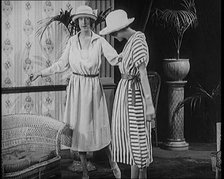 Two Female Civilians Leisuring in a Drawing Room Wearing Long Dresses and Hats Comparing..., 1920. Creator: British Pathe Ltd.