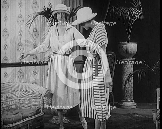 Two Female Civilians Leisuring in a Drawing Room Wearing Long Dresses and Hats Comparing..., 1920. Creator: British Pathe Ltd.