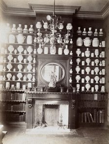 Drug jars and books on shelves surrounding a mantlepiece with a statue and a mirror, c1900.. Creator: Unknown.