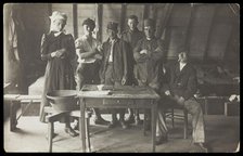 French prisoners of war, one in drag, gather round a small wooden table, [between 1910 and 1919?]. Creator: Unknown.