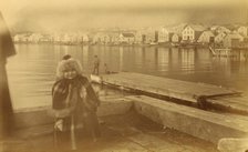 Unidentified young girl standing on wharf, with buildings along waterfront...,1894 or 1895. Creator: Alfred Lee Broadbent.