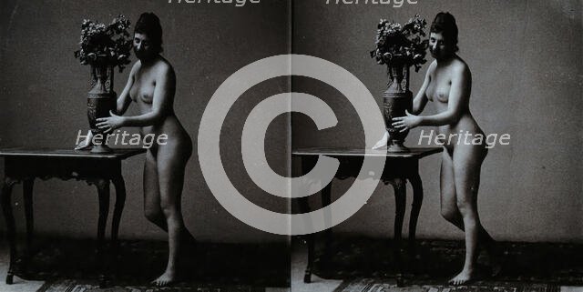 A young woman posing naked in a photographic studio, standing in front of a table on which..., c1900 Creator: Unknown.