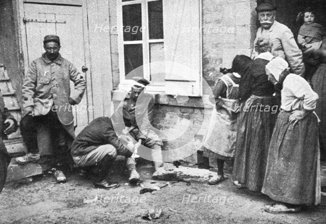 'Derriere L'ennemi refoule; Un major soignant un allemand blesse que des villageois..., 1915 (1924). Creator: Unknown.