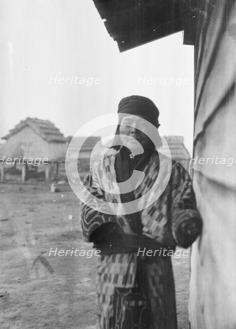 Ainu standing outside by the wall of a wooden hut, 1908. Creator: Arnold Genthe.