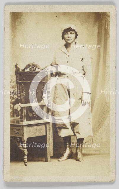 Photographic postcard of a woman standing next to a wooden chair, 1918-1930. Creator: Unknown.