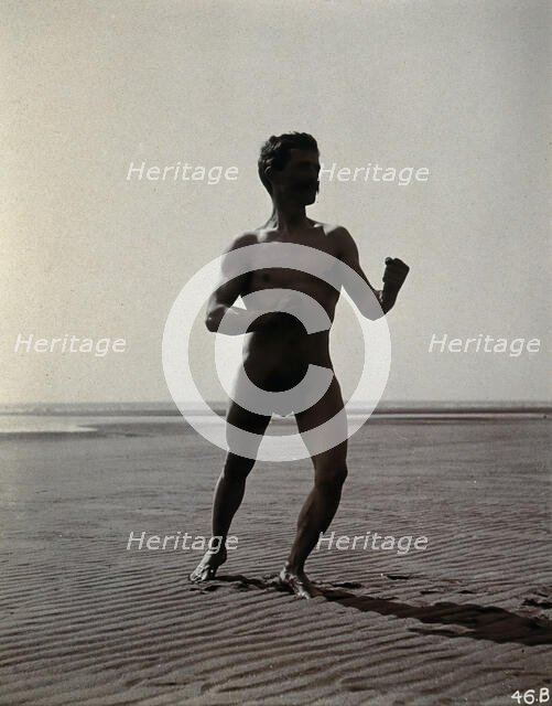 A man posing naked in a boxing stance with his fists raised, on a deserted beach, c1900. Creator: Unknown.