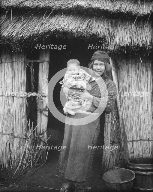 Ainu woman holding a child standing outside a hut, 1908. Creator: Arnold Genthe.