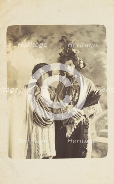Two men in drag, one wearing a detailed feathered head garment, pose for a portrait, [1900-1909?]. Creator: Frank Wood.