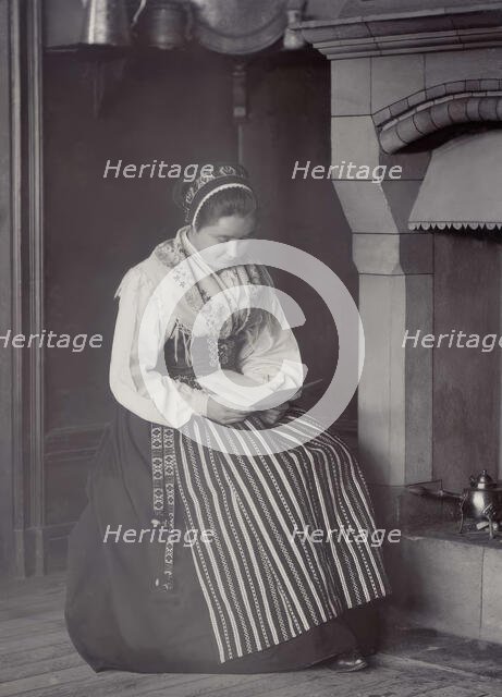 A woman sits in front of a fireplace and reads dressed in a Dalarna costume, 1886-1920. Creator: Helene Edlund.