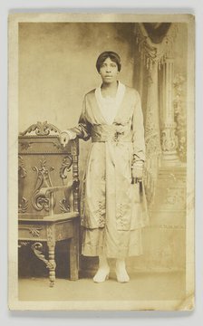 Photographic postcard of a woman standing next to a wooden chair, 1904-1918. Creator: Unknown.
