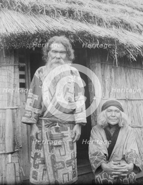 Ainu man and seated woman at the entrance of a hut, 1908. Creator: Arnold Genthe.