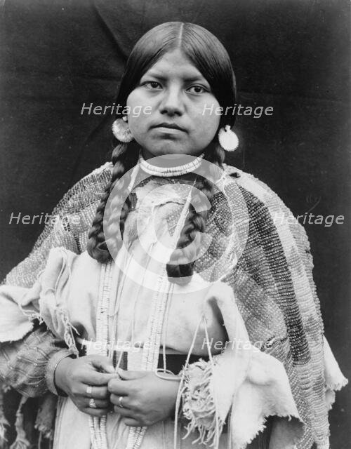 Cayuse woman, half-length portrait, standing, facing front, braids, shell disk earrings..., c1910. Creator: Edward Sheriff Curtis.
