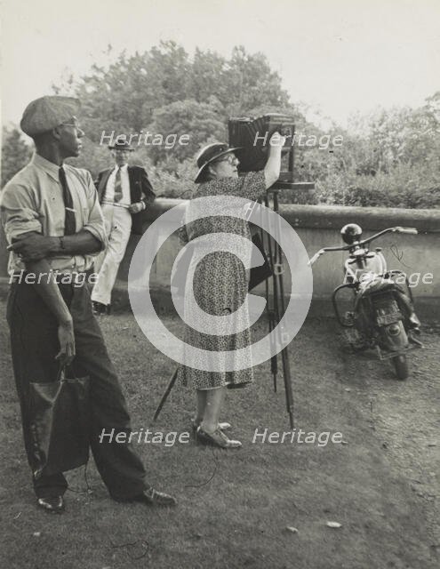 Photographer Frances Benjamin Johnston standing beside her view camera preparing to take..., 1938. Creator: Unknown.