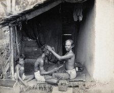 Two men sitting cross-legged; one is a barber shaving the front of the other's head, c1900. Creator: Unknown.