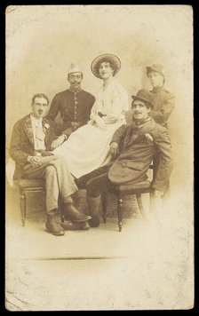 Amateur actors sitting down, surrounding a man in drag seated in the centre, [1910-1919?]. Creator: Unknown.