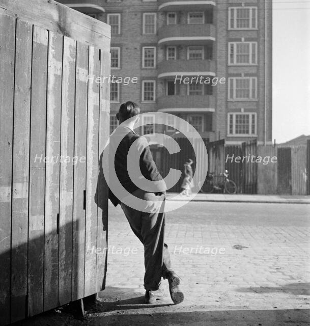 A man leaning on a street corner hoarding, possibly Rugmere on Ferdinand St, Camden, 1945-50. Creator: John Gay.
