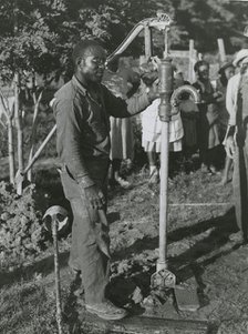 An African American man fixing a well's tube into the ground near Ridge, Maryland, July 1941. Creators: Farm Security Administration, John Collier.