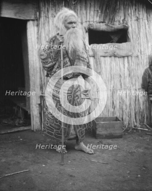 Ainu man holding a staff standing outside a hut, 1908. Creator: Arnold Genthe.