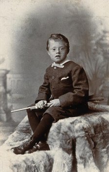 A boy with Down's syndrome, holding a rifle and sitting on a fur carpet, c1900. Creator: RW Morris.