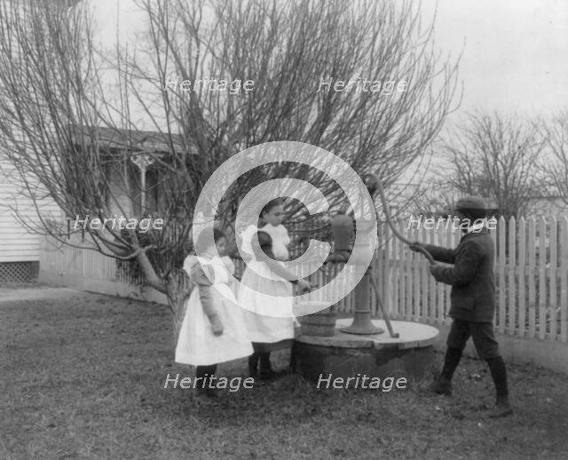 Two girls and a boy pumping water at well of Hampton Institute graduate, 1899 or 1900. Creator: Frances Benjamin Johnston.