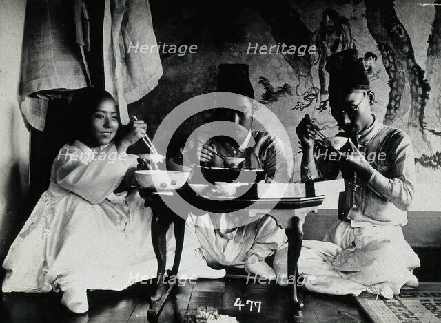 A group of people eating a meal, seated behind a small table, in front of a painted screen, c1900. Creator: Unknown.