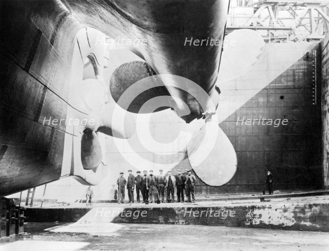 Workmen standing under one of the propellors of the Titanic, 31 May, 1911 (b/w photo)