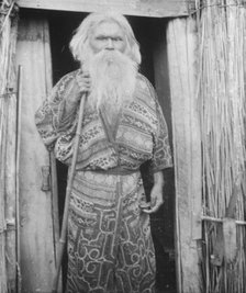 Ainu man holding a staff standing at the doorway of a hut, 1908. Creator: Arnold Genthe.