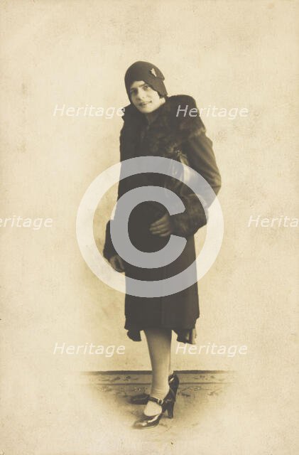 A person poses wearing a long coat with fur trim and a hat with a brooch, [between 1920 and 1929?]. Creator: Unknown.