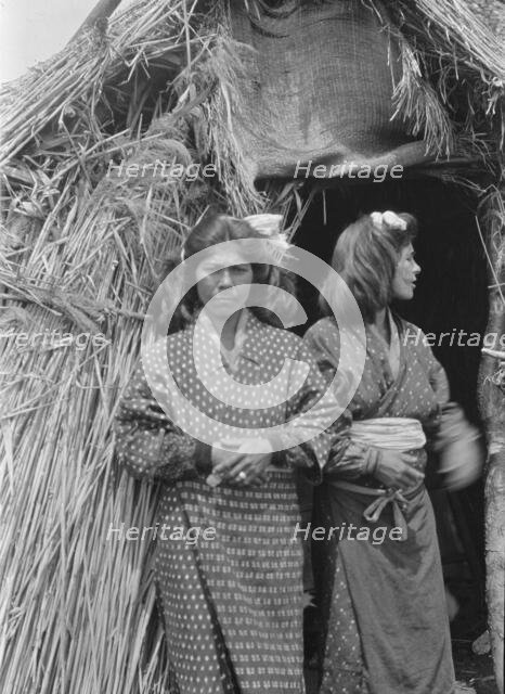 Two Ainu women standing outside the entrance of a hut, 1908. Creator: Arnold Genthe.
