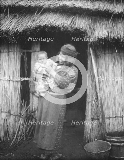 Ainu woman holding a child standing outside a hut, 1908. Creator: Arnold Genthe.