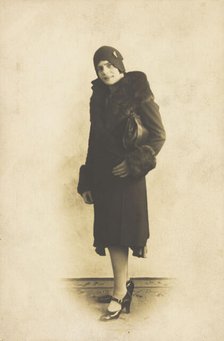 A person poses wearing a long coat with fur trim and a hat with a brooch, [between 1920 and 1929?]. Creator: Unknown.