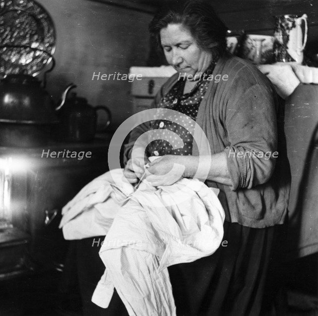 A woman sewing a shirt by the stove, London, 1953. Artist: Henry Grant