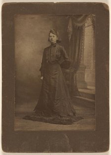 Portrait of a young woman in long black dress, standing, about 1900. Creator: George W Killian.